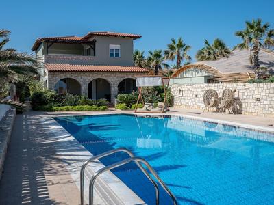 Gran piscina exterior con agua azul frente a una casa de dos pisos con arcadas y palmeras.