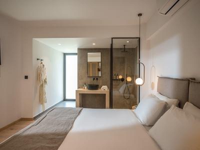 Habitación de hotel moderna con cama grande, pared de vidrio al baño y diseño minimalista.