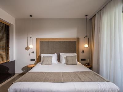 Jasny pokój hotelowy z łóżkiem podwójnym, beżową pościelą i nowoczesnymi lampami wiszącymi po obu stronach.