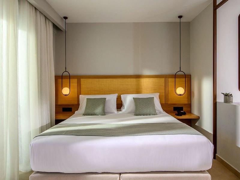 Habitation de hotel luminosa con cama grande, dos almohadas y ventanas de piso a techo con cortinas blancas.