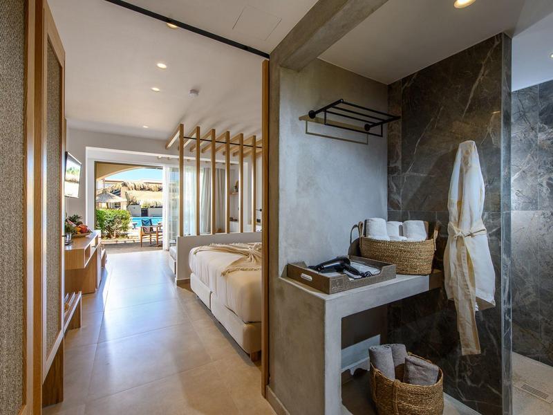 Habitación de hotel moderna con cama, baño abierto y vista a una terraza con piscina.