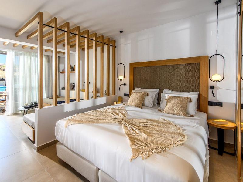 Habitación de hotel luminosa con cama grande, detalles de madera y vista al mar a través de una pared de vidrio.