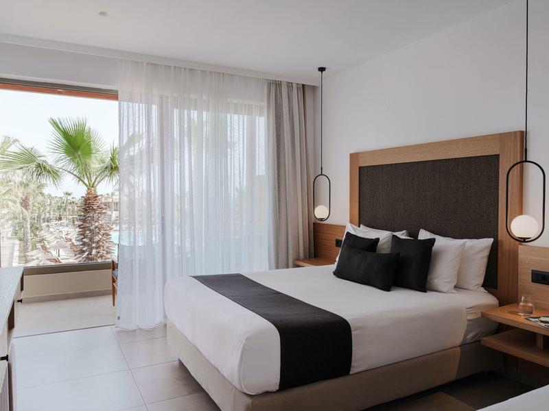 Dormitorio moderno con cama grande, acentos negros y vista de palmeras a través de la ventana.