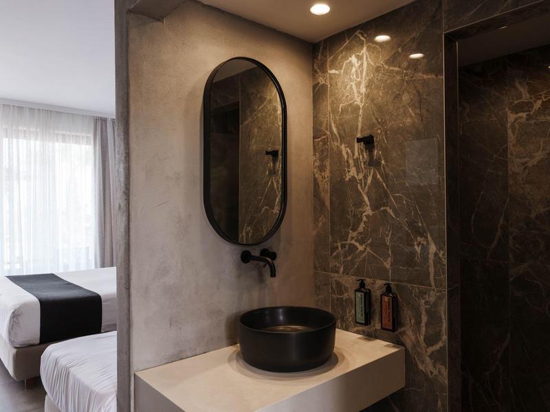 Baño moderno con lavabo negro, espejo y pared de mármol junto a un dormitorio luminoso.