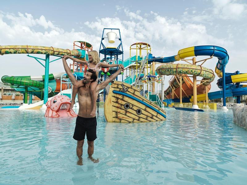 Hombre de pie en agua poco profunda frente a un parque acuático colorido con toboganes y estructuras de juego.