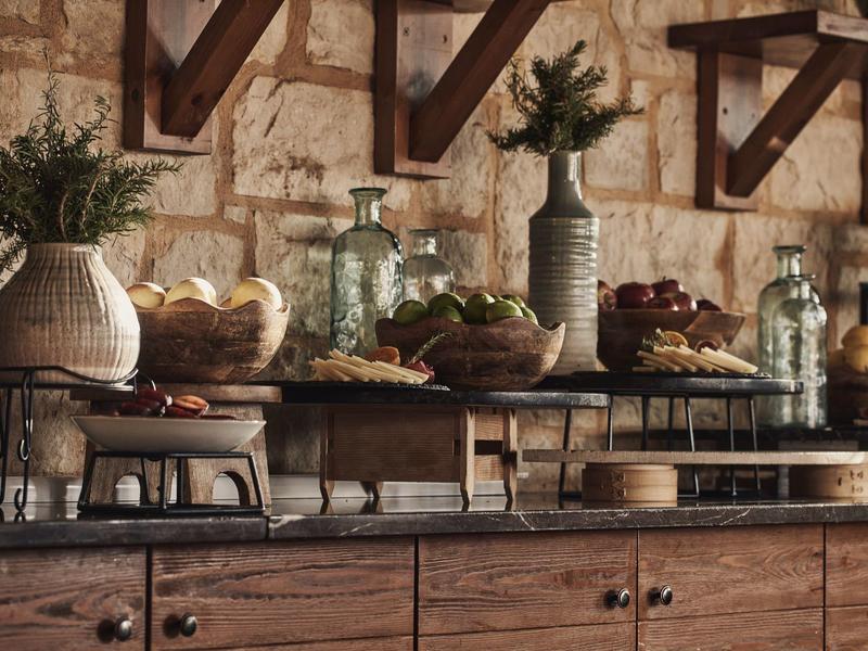 Mostrador de desayuno rústico con cestas, flores y dispensadores de bebidas frente a pared de piedra.