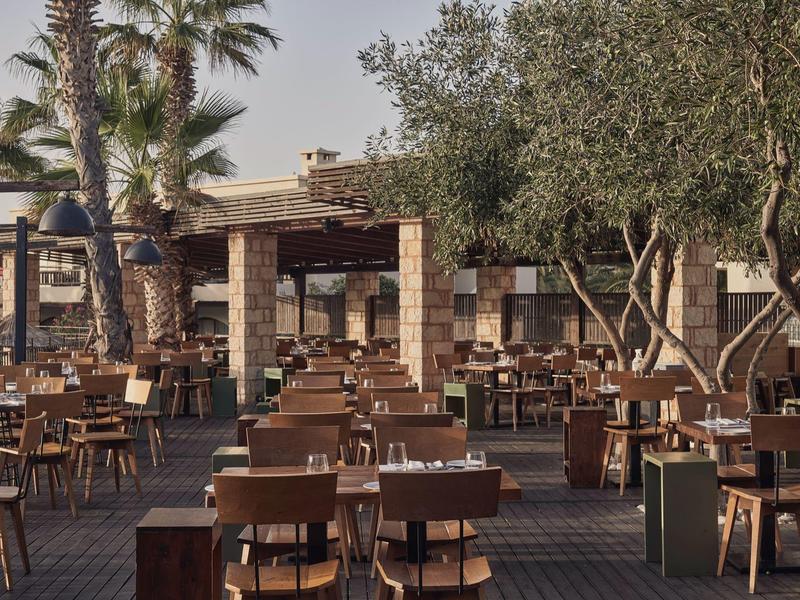 Restaurante al aire libre con sillas y mesas de madera bajo palmeras y otros árboles.