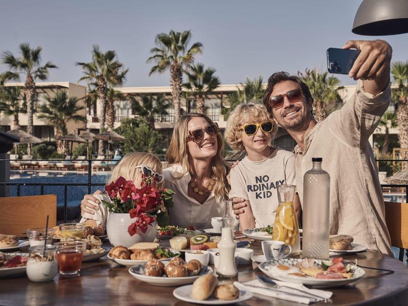 Tres personas se toman una selfie en una mesa de desayuno en un hotel con piscina y palmeras al fondo.