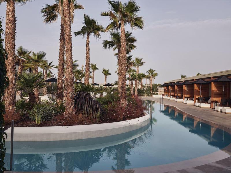 Una piscina de hotel tranquila con palmeras y áreas de descanso sombreadas bajo un cielo nublado.