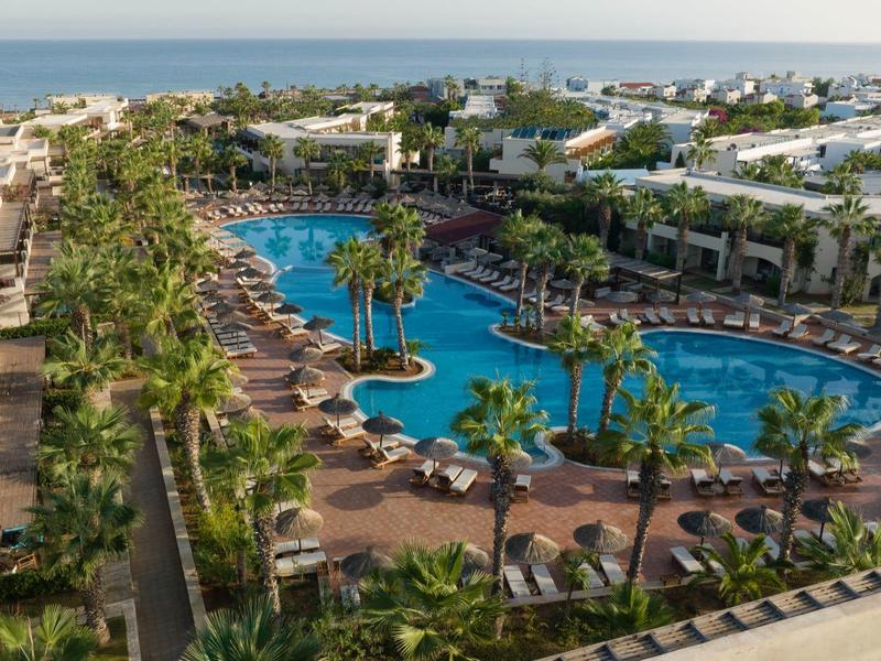 Vista de un resort hotelero con una gran piscina, palmeras y tumbonas junto al mar.