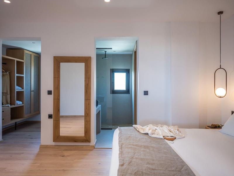 Dormitorio luminoso con cama grande, espejo de pared y baño abierto con ventana.