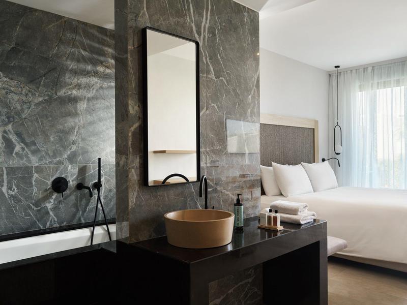 Habitación de hotel moderna con cama, gran espejo, lavabo y bañera con azulejos oscuros.