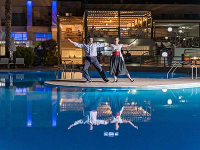 Zwei Personen tanzen bei Nacht neben einem beleuchteten Pool vor einem Gebäude mit großen Fenstern.