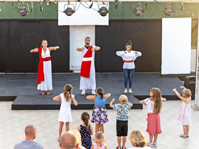 Kinder lernen auf Bühne mit Erwachsenen Tanzschritte, weiße und rote Kleidung, sonniger Tag.
