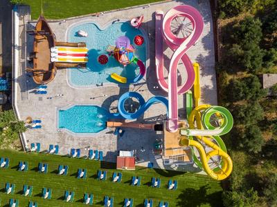 Wasserpark mit bunten Wasserrutschen, Schwimmbecken und Liegestühlen in grüner Umgebung.