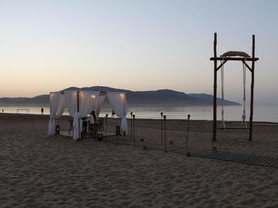 Zelt mit weißen Vorhängen und Holzgestell am Strand bei Dämmerung, Hintergrund mit Bergen und ruhigem Wasser.