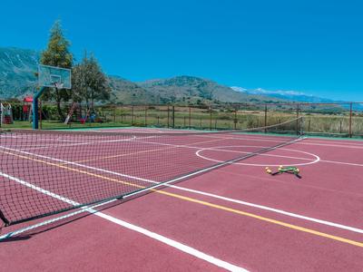 Tennisplatz mit rotem Boden, Netz, umgeben von grüner Landschaft und Bergen im Hintergrund.
