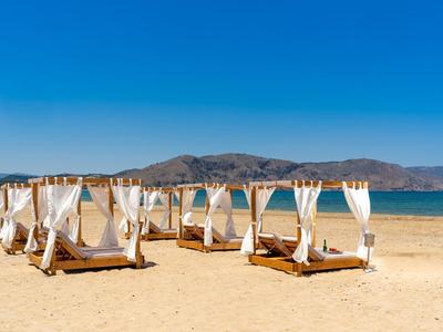 Strand mit weißen, hölzernen Tagesbetten mit Vorhängen und blauem Himmel im Hintergrund.