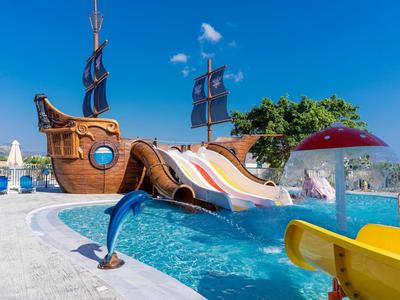 Bunter Wasserspielplatz mit Piratenschiffrutsche, Fontänen und klarem blauem Wasser unter blauem Himmel.