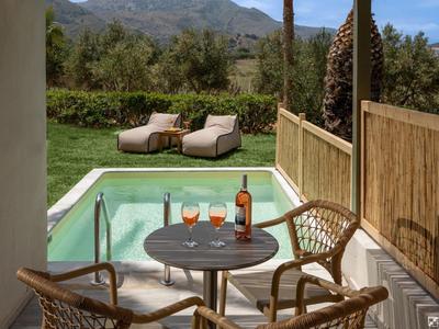 Terrasse mit Holztisch, zwei Stühlen, Pool, zwei Liegestühlen und grüner Landschaft im Hintergrund.