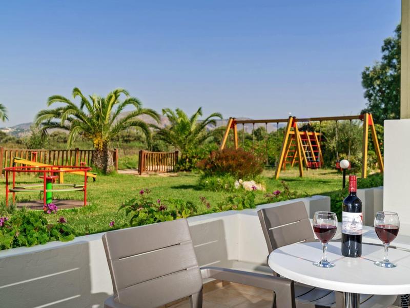 Terrace with table, wine, and view of a green garden with swings and palm trees.