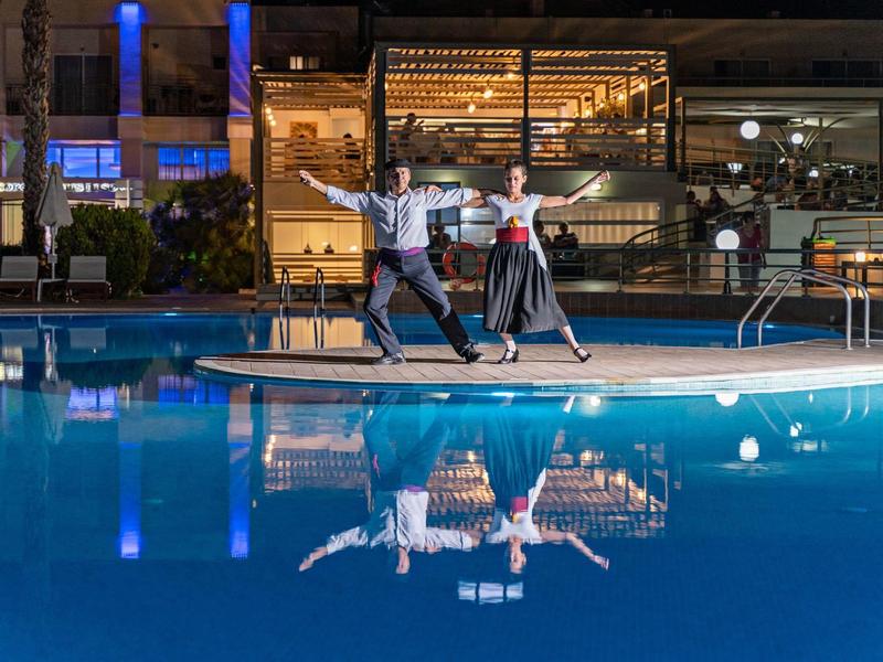 Couple dancing by the lit hotel pool at night with reflection in the water.