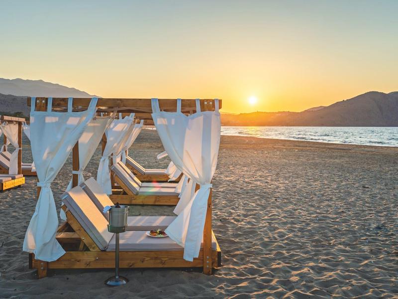 Beach loungers with white curtains on sandy beach at sunset with ocean in the background.