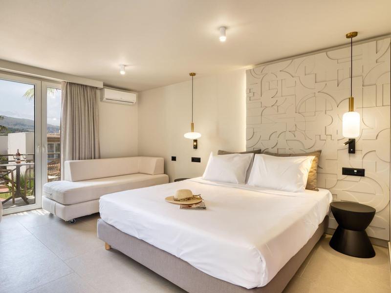 Bright hotel room with king bed, sofa, and balcony overlooking mountains.