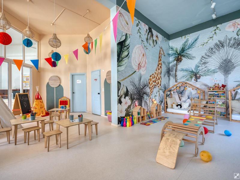 Colorful kindergarten room with tables, chairs, and a wall mural of animals and plants.