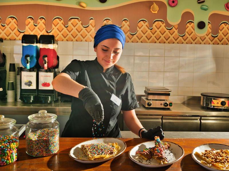 Chef sprinkles toppings on waffles in a kitchen with colorful candies and waffle makers.