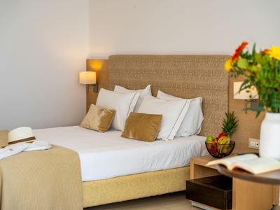 Hell eingerichtetes Schlafzimmer mit großem Bett, Kissen, Nachttisch, Lampe und buntem Blumenstrauß.