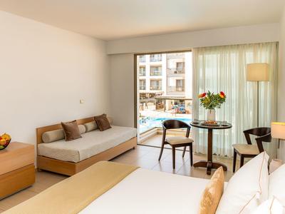 Helles Zimmer mit großem Bett, Sofa, Tisch, Stühlen und Balkon mit Blick auf Pool und Gebäude.