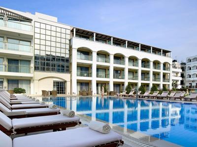 Moderní hotelový bazén s lehátky a světlou budovou v pozadí pod jasnou oblohou.