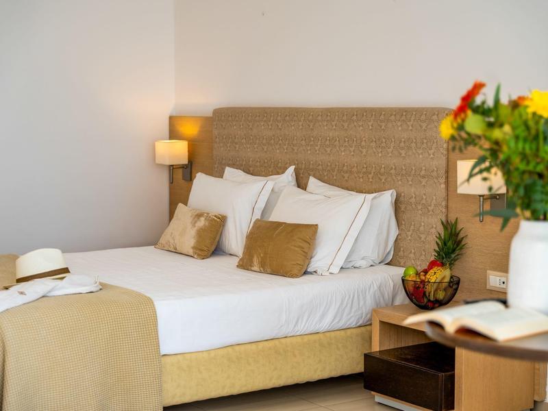 Przytulny pokój hotelowy z podwójnym łóżkiem, beżowymi poduszkami, lampą i kolorowymi kwiatami.