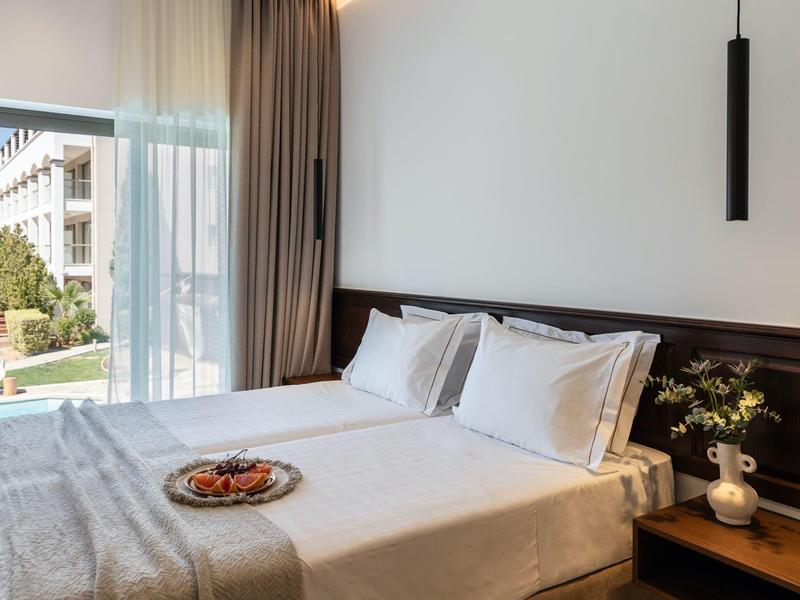 Jasny pokój hotelowy z podwójnym łóżkiem, balkonem i widokiem na zielony ogród.