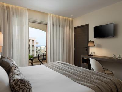 Modernes Hotelzimmer mit großem Fenster, Doppelbett, Schreibtisch und TV, Blick auf Balkon und Gebäude.