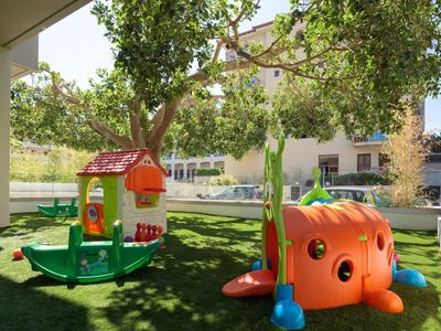 Kinderspielplatz mit grünem Rasen, Spielhaus und Spielgeräten im Hotelgarten bei Sonnenschein.
