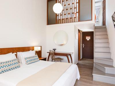 Helles Hotelzimmer mit Doppelbett, Schreibtisch, Spiegel, Treppe und Fernseher an der Wand.