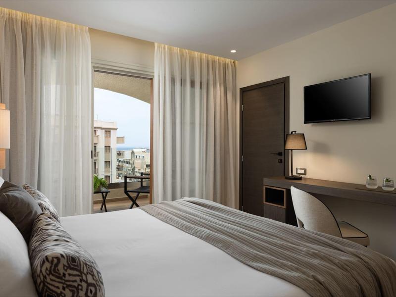 Modernes Hotelzimmer mit großem Bett, Schreibtisch, Fernseher und Balkon mit Blick auf Gebäude.