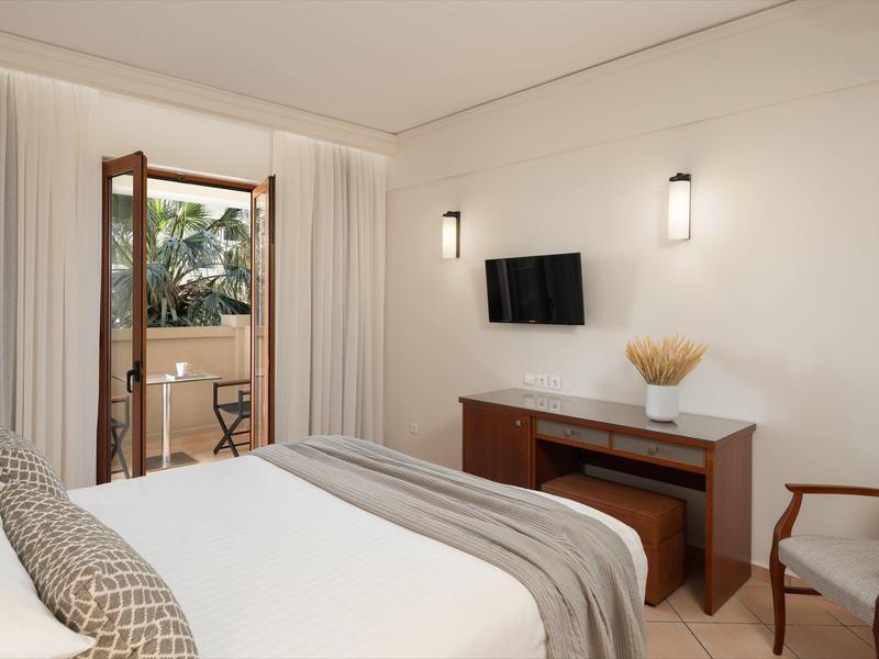 Modernes Hotelzimmer mit Bett, Schreibtisch, Stuhl und Blick auf den Balkon mit Palmen.