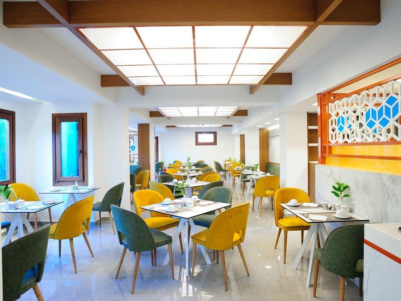 Heller, moderner Speiseraum mit gelben und grünen Stühlen, weißen Tischen und großem Deckenlicht.