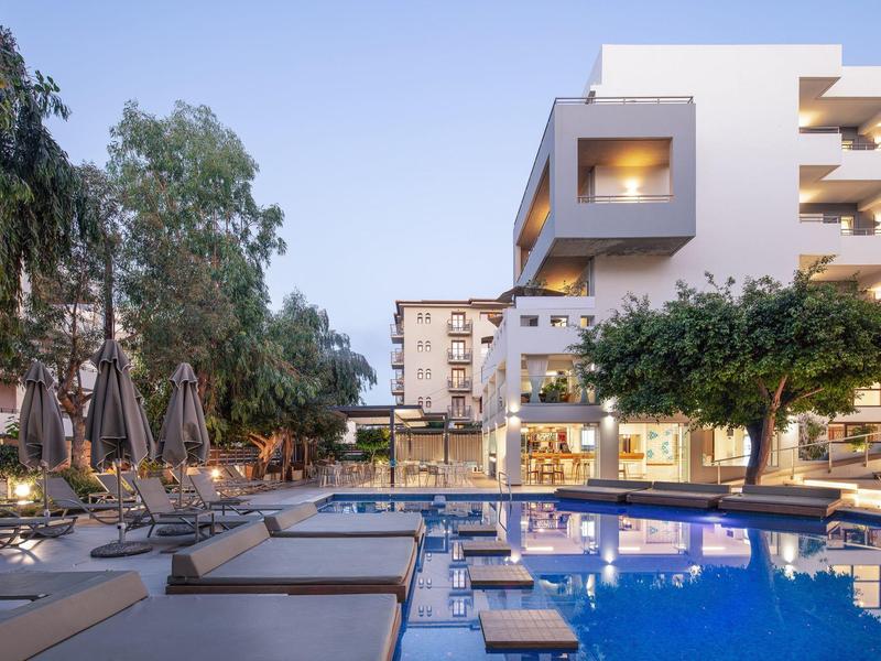 Moderner Hotelpool bei Dämmerung mit Liegen, Sonnenschirmen und Gebäuden im Hintergrund.