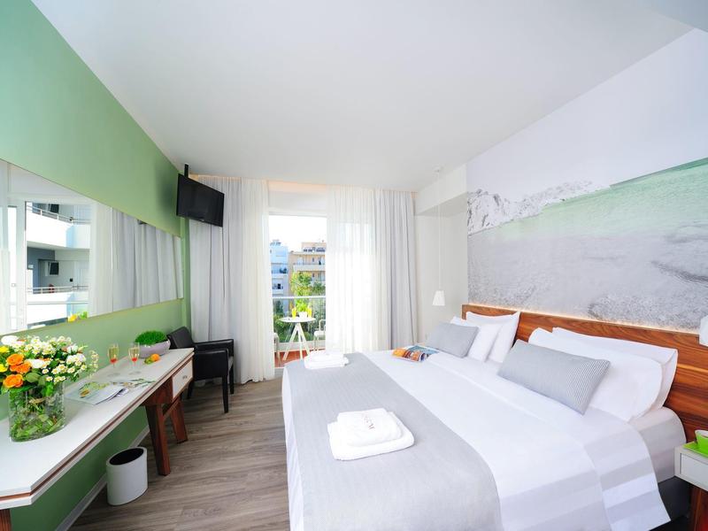 Modernes Hotelzimmer mit großem Bett, grün-weißer Wand, Fernseher und Schreibtisch mit Blumen.