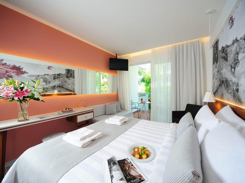 Modernes Hotelzimmer mit großem Bett, weißer Bettwäsche, Blumenstrauß und einem Fernseher an der Wand.