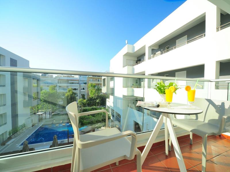 Balkon mit weißem Tisch und zwei Stühlen, Glasgeländer, Blick auf Pool und moderne weiße Gebäude.