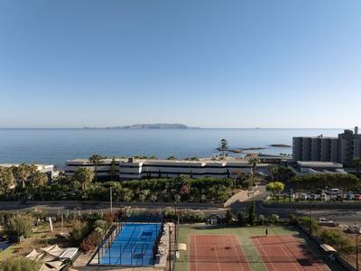 Vista sul mare, marina turistico e campi da tennis accanto a un complesso alberghiero sotto un cielo sereno.