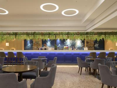Area lounge moderna dell'hotel con bar blu, sedie grigie e luci LED circolari sul soffitto.