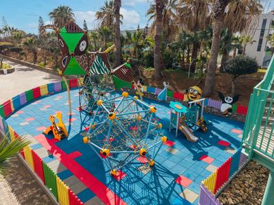 Parco giochi per bambini con strutture colorate per arrampicarsi e scivolo, circondato da palme e edifici.