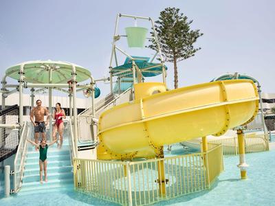 Kinder spielen in einem klaren blauen Pool mit einer gelben Wasserrutsche und einer grünen Plattform.