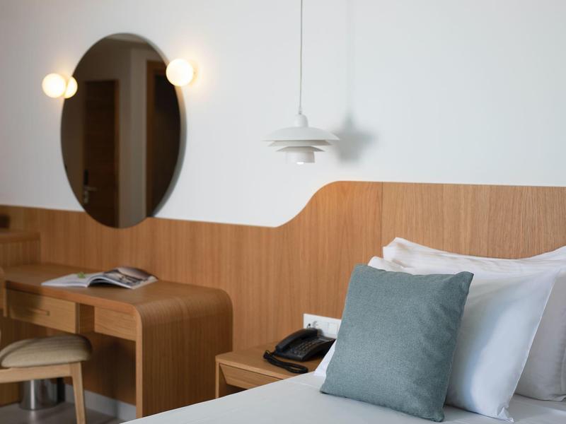 Hotelový pokoj s bílou postelí, šedým polštářem, oválným zrcadlem a dřevěným stolem.